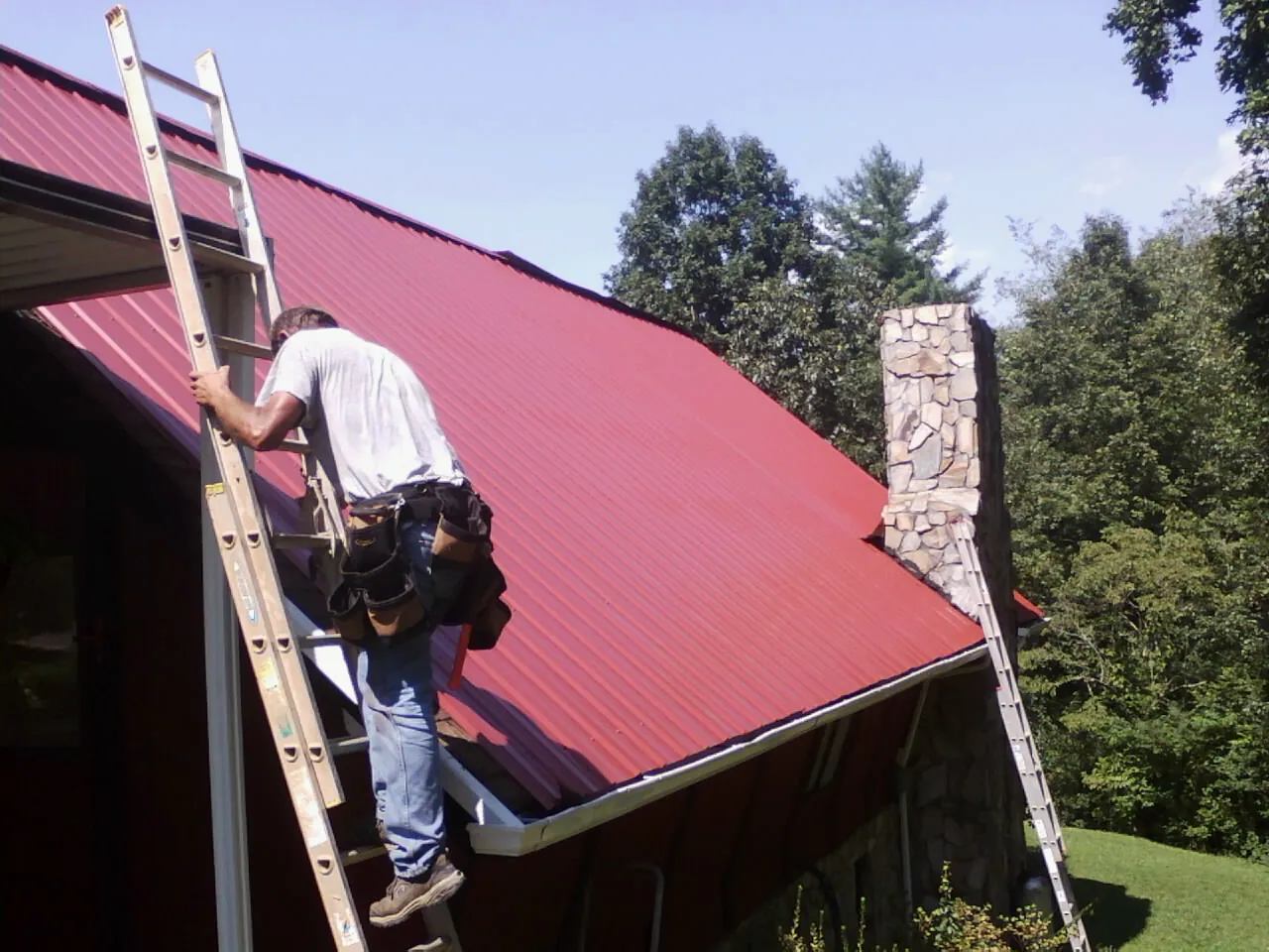 Roofer on a ladder beside a red metal roof and stone chimney — Soffit & Fascia Repair by Roofing Brothers in Benton
